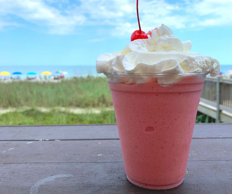 Pink frozen drink with whipped cream sitting on table with ocean in the background