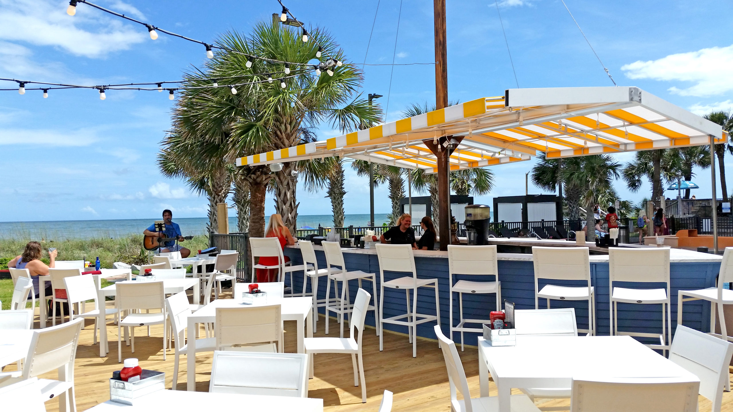 An outdoor bar with white tables and chairs, a blue and yellow striped awning, and a view of the ocean.